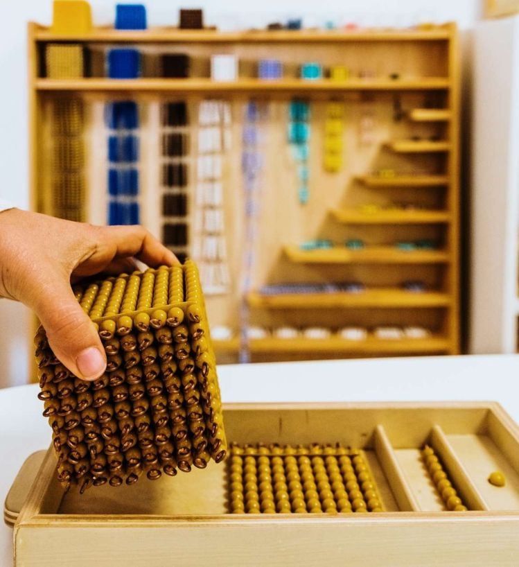 A guide's hand lifting the golden bead thousand cube from its tray, in front of a shelf of Montessori mathematics materials